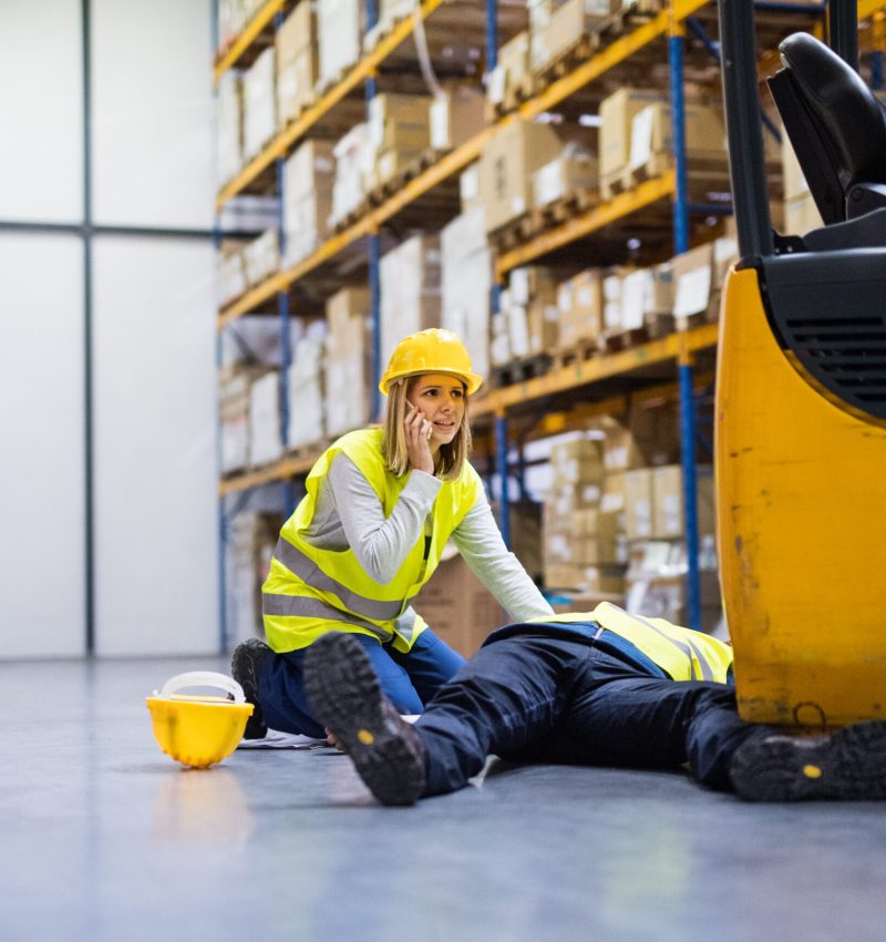 An accident in a warehouse. Woman with smartphone and her colleague lying on the floor next to a forklift.